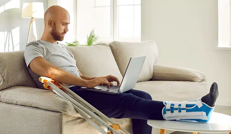 Unhealthy,Businessman,With,Injured,Leg,In,Bandage,Sit,On,Sofa Unhealthy businessman with injured leg in bandage sit on sofa at home work online on computer. Unwell male employee with foot trauma wearing special splint use laptop. Injury and rehabilitation.